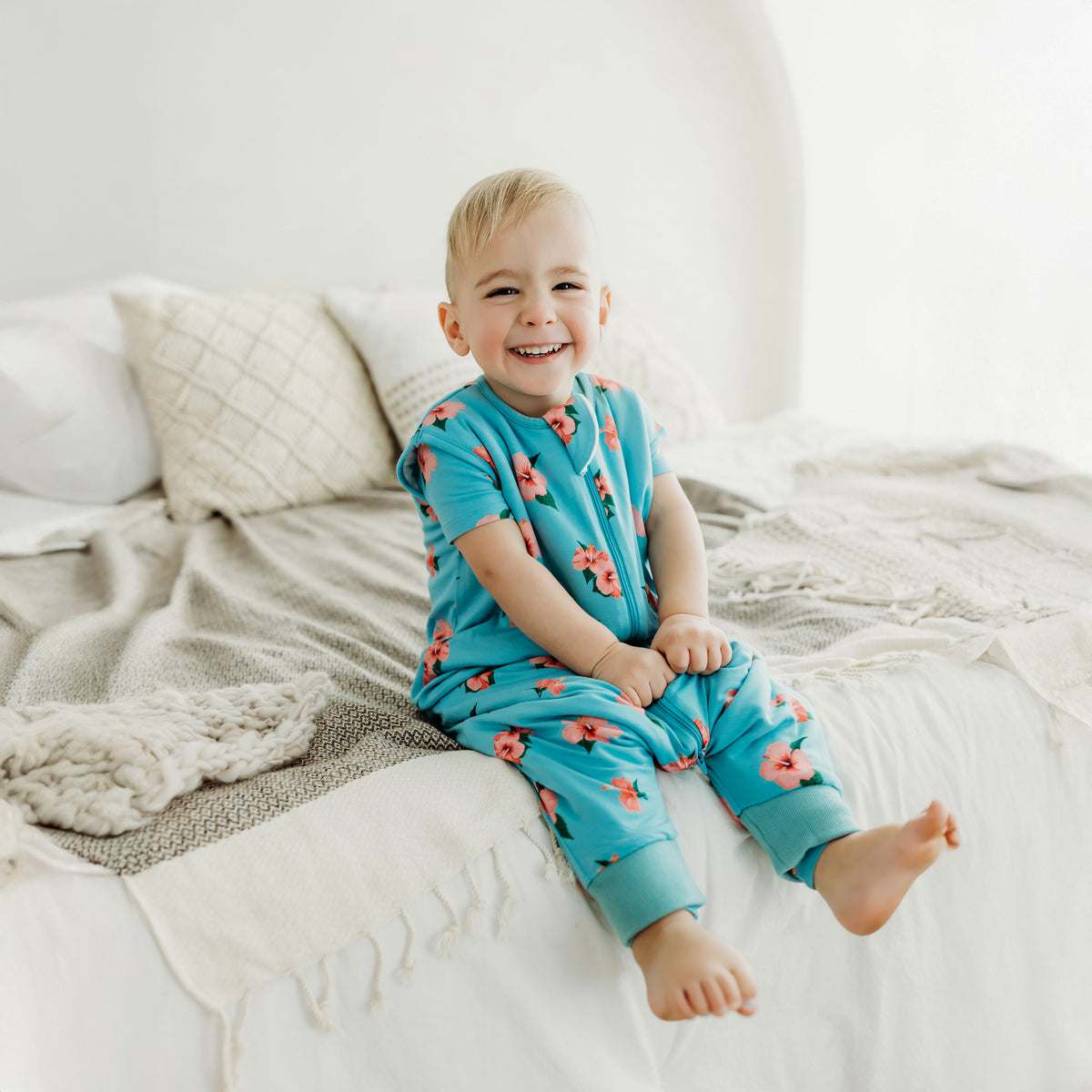 Smiling toddler sitting on a bed wearing the Sleep Bag Walker in Hibiscus 1.0
