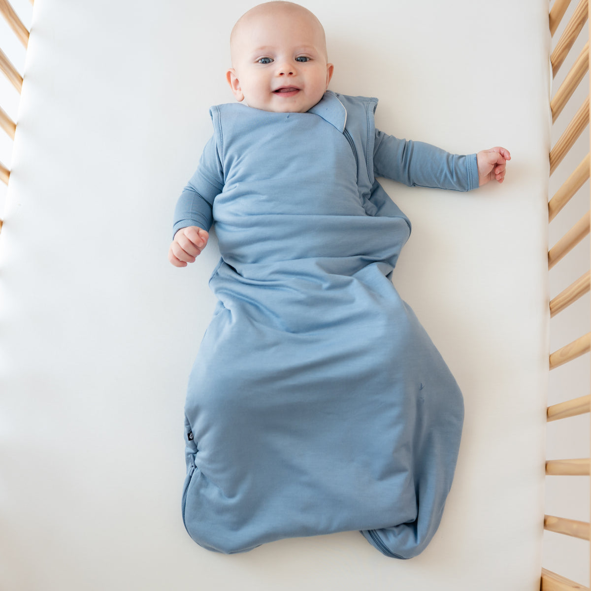 Baby laying in crib wearing Kyte Baby Sleep Bag in Slate 1.0 with matching bodysuit underneath