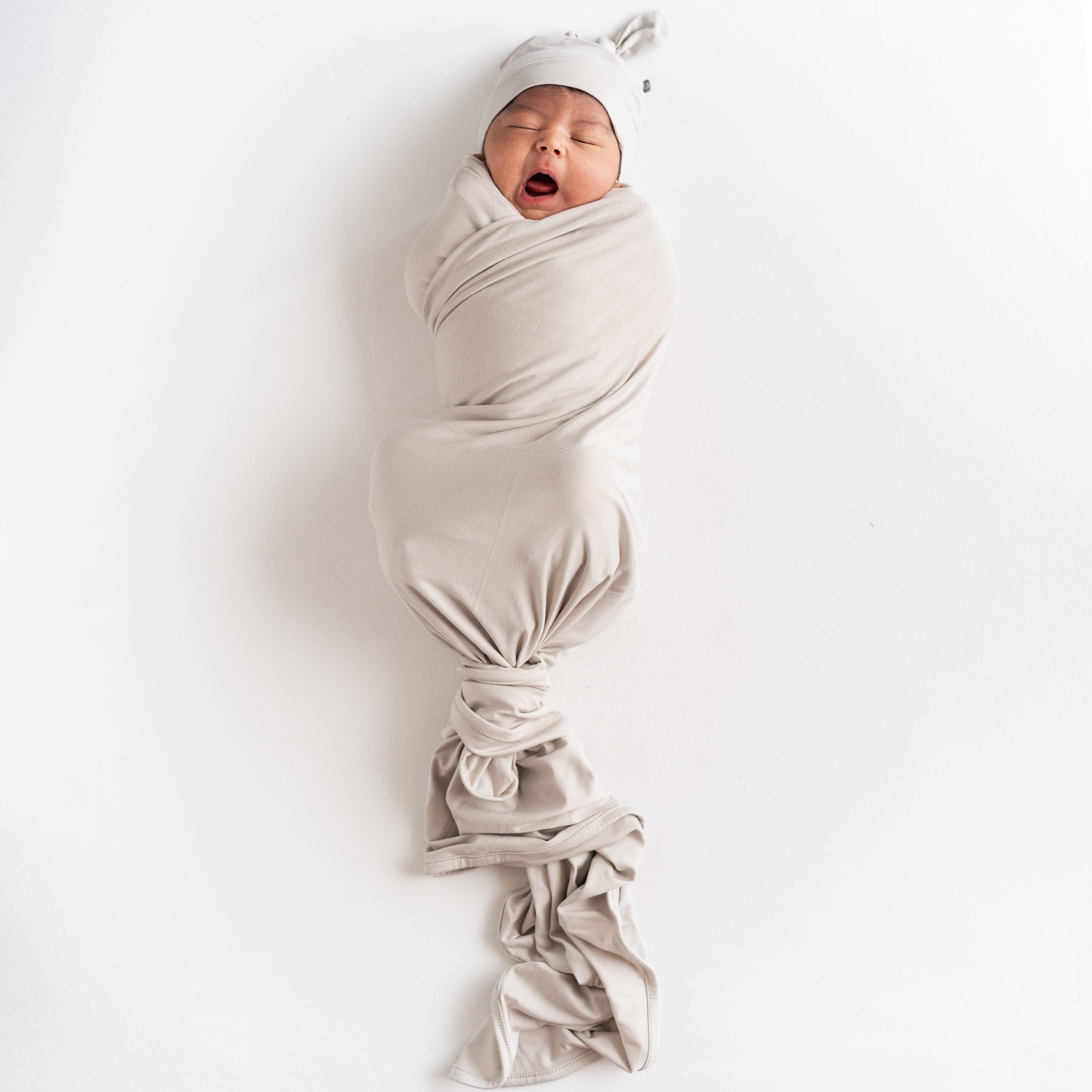 Yawning infant wrapped in a swaddle in Oat and matching knotted cap