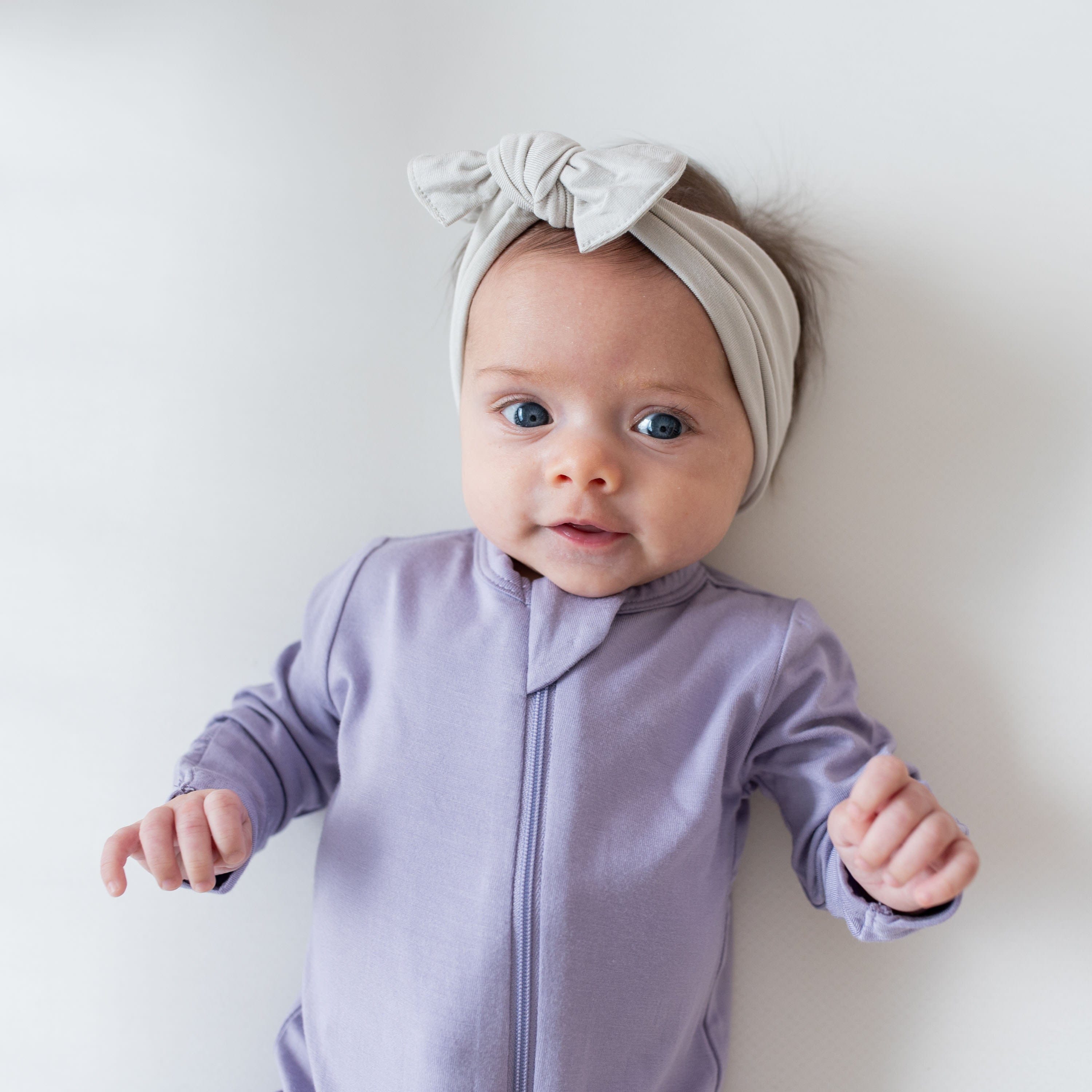 Infant wearing a Zippered Footie in Taro and a headband bow in Oat
