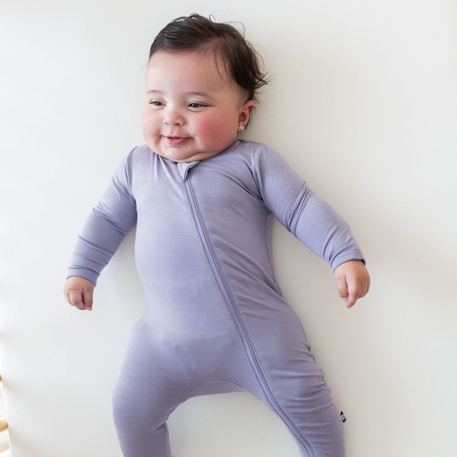Smiling baby wearing a Zippered Footie in Taro while laying on an off-white surface
