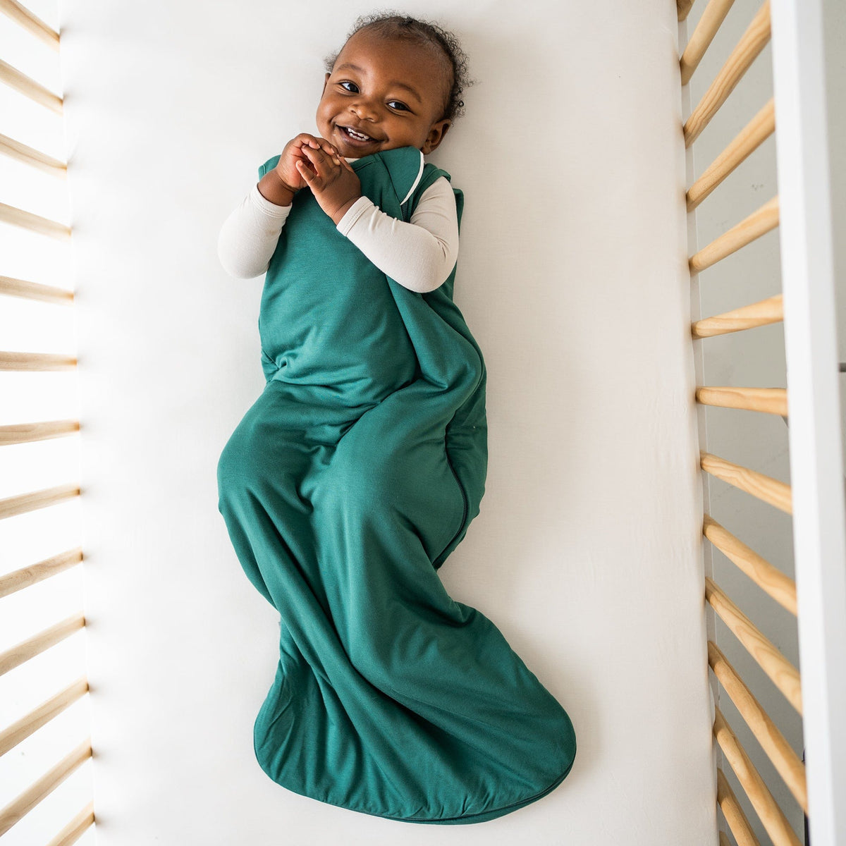 Smiling baby laying in a crib while wearing Kyte Baby Sleep Bag in Emerald 2.5 with an Oat long sleeve romper underneath